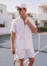 Man in white outfit with cap and sunglasses on a tennis court