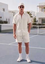 Man in a white outfit standing on a tennis court with modern buildings and palm trees in the background.