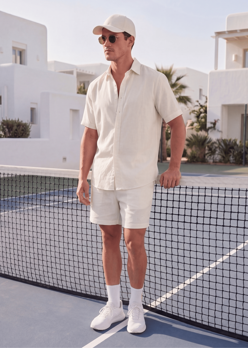 Man in a white outfit standing on a tennis court with white buildings and palm trees in the background.