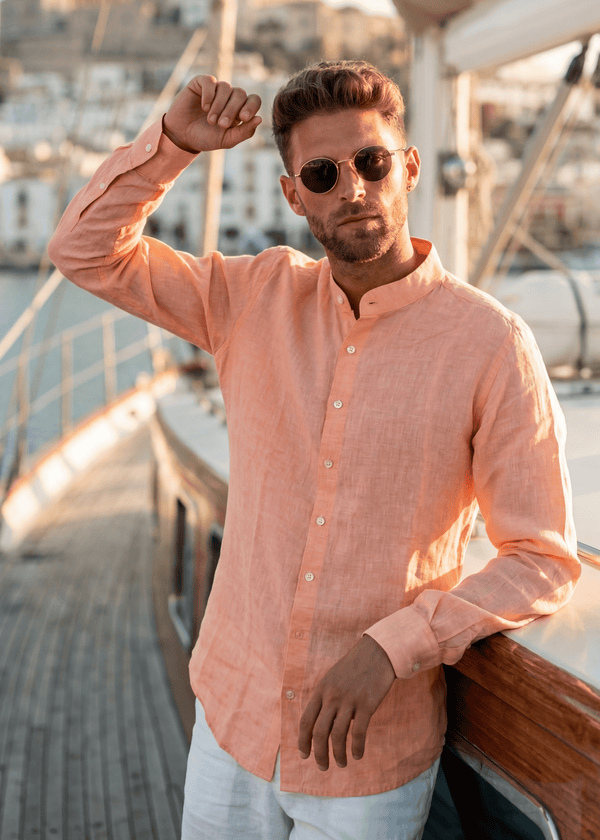 PIETER PETROS PP Male linen shirt in orange, Chinese collar, seashell buttons, worn on a sailboat deck overlooking a Mediterranean harbour at golden hour