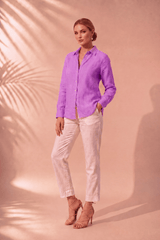 A woman stands in a studio wearing a bright lavender-purple long-sleeve button-up shirt and off-white cropped pants, paired with nude high-heel sandals. The background is a warm peach-pink studio backdrop with soft palm leaf shadows.