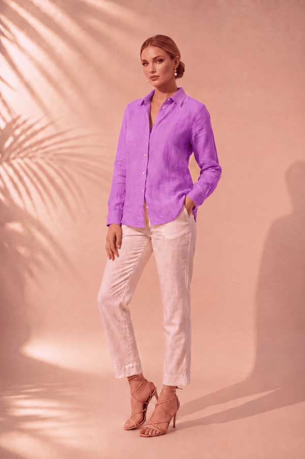 A woman stands in a studio wearing a bright lavender-purple long-sleeve button-up shirt and off-white cropped pants, paired with nude high-heel sandals. The background is a warm peach-pink studio backdrop with soft palm leaf shadows.
